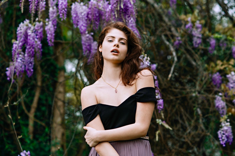 May include: A woman with long brown hair wearing a black off-the-shoulder top and a purple skirt stands in front of a wall of purple wisteria flowers.