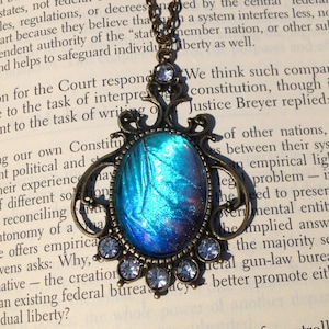 Puede incluir: Un collar con un colgante de bronce con un dise&ntilde;o de ala de mariposa iridiscente azul y p&uacute;rpura. El colgante est&aacute; rodeado de diamantes de imitaci&oacute;n transparentes. El collar est&aacute; sobre un libro con texto sobre la constituci&oacute;n.