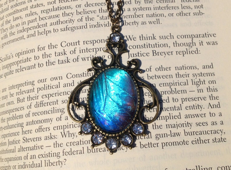 Puede incluir: Un collar con un colgante de bronce con un dise&ntilde;o de ala de mariposa iridiscente azul y p&uacute;rpura. El colgante est&aacute; rodeado de diamantes de imitaci&oacute;n transparentes. El collar est&aacute; sobre un libro con texto sobre la constituci&oacute;n.