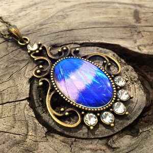 Op de afbeelding: Een vintage-stijl hangerketting met een ovale, iriserende blauwe en paarse middensteen. De hanger is omlijst door een sierlijke, bronskleurige metalen filigraan en geaccentueerd met heldere, ronde kristallen. De ketting is ook bronskleurig.