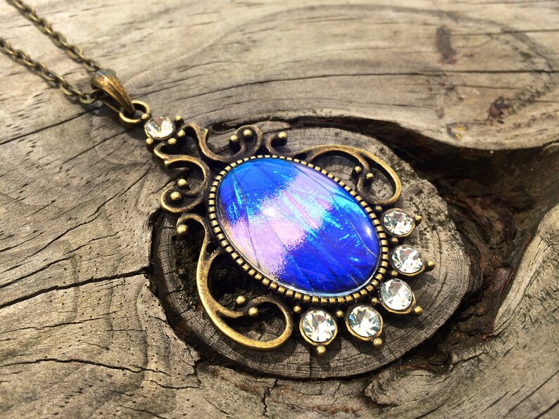 Op de afbeelding: Een vintage-stijl hangerketting met een ovale, iriserende blauwe en paarse middensteen. De hanger is omlijst door een sierlijke, bronskleurige metalen filigraan en geaccentueerd met heldere, ronde kristallen. De ketting is ook bronskleurig.