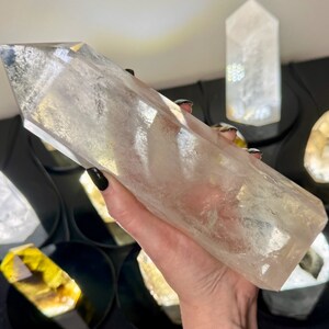 May include: A large, clear quartz crystal point, also known as a crystal wand, is held in a hand. The crystal is smooth and polished, with a pointed tip and a flat base. The crystal is surrounded by other smaller crystals, including a yellow citrine crystal.