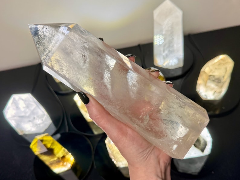 May include: A large, clear quartz crystal point, also known as a crystal wand, is held in a hand. The crystal is smooth and polished, with a pointed tip and a flat base. The crystal is surrounded by other smaller crystals, including a yellow citrine crystal.