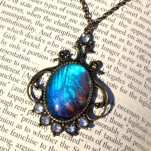 Puede incluir: Un collar con colgante de bronce con un ala de mariposa azul y morada encapsulada en resina. El colgante est&aacute; adornado con brillantes transparentes y cuelga de una cadena delicada.