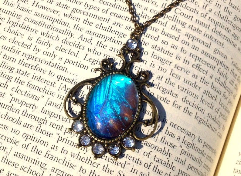 Puede incluir: Un collar con colgante de bronce con un ala de mariposa azul y morada encapsulada en resina. El colgante est&aacute; adornado con brillantes transparentes y cuelga de una cadena delicada.