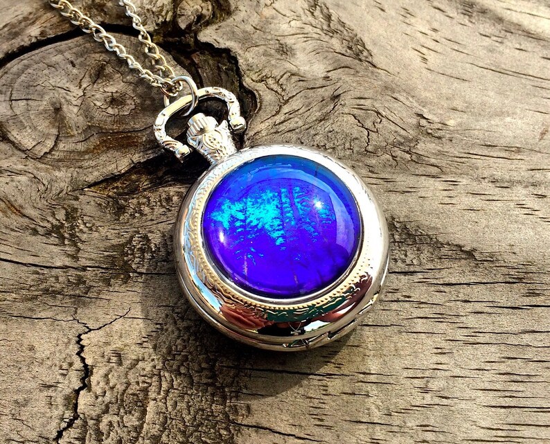 Op de afbeelding: Een zilveren zakhorlogeketting met een blauw en groen iriserend glazen gezicht.
