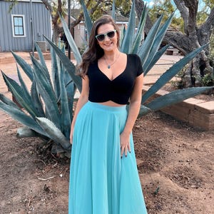 A woman in a black crop top and a long turquoise skirt stands in front of a large blue agave plant, smiling and posing for the camera.