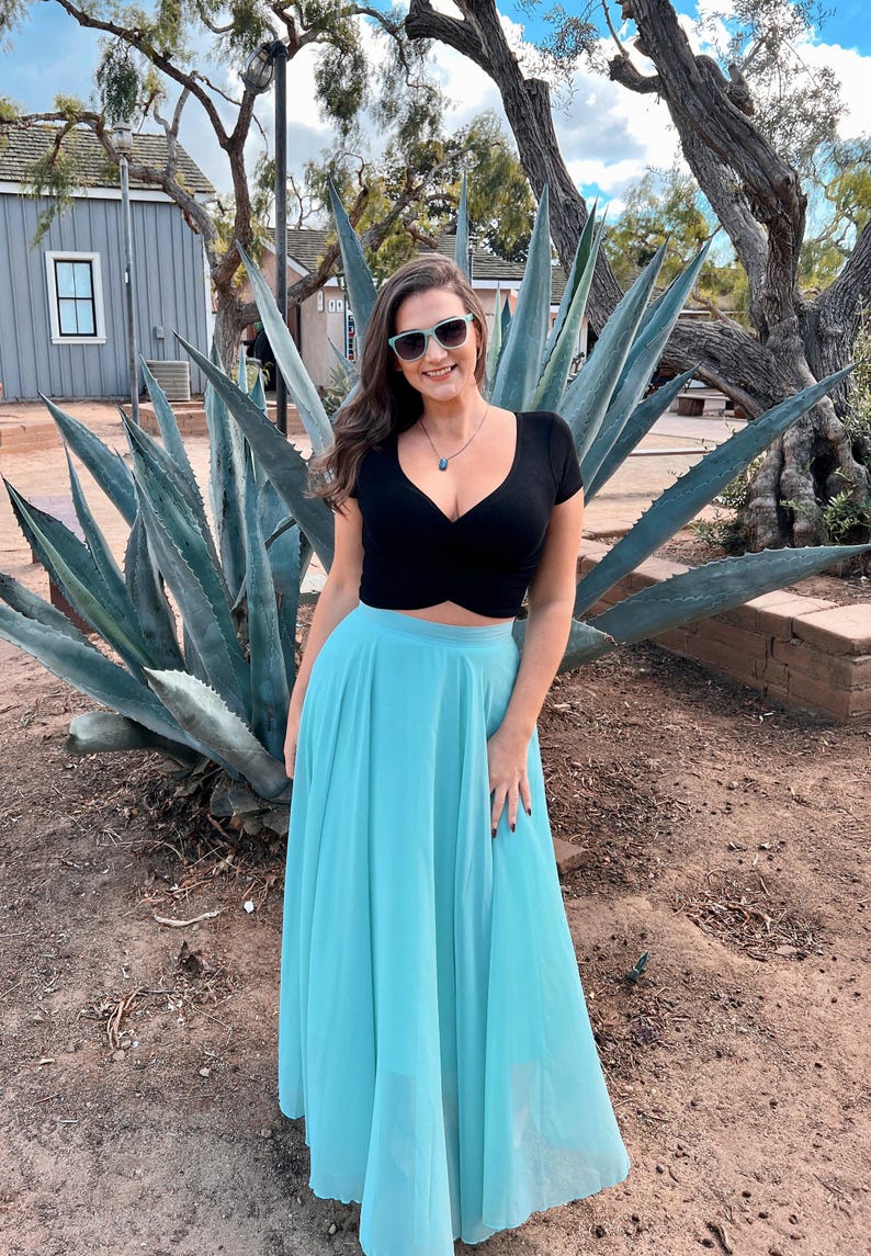 A woman in a black crop top and a long turquoise skirt stands in front of a large blue agave plant, smiling and posing for the camera.