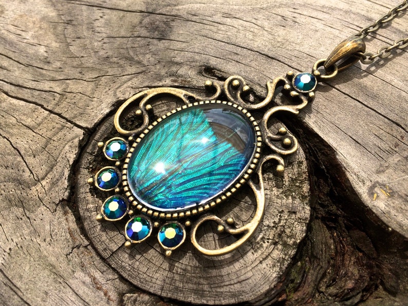 May include: A gold-tone pendant necklace featuring a large oval-shaped blue and green iridescent glass cabochon set in a filigree frame. The pendant is surrounded by smaller blue and green rhinestones.
