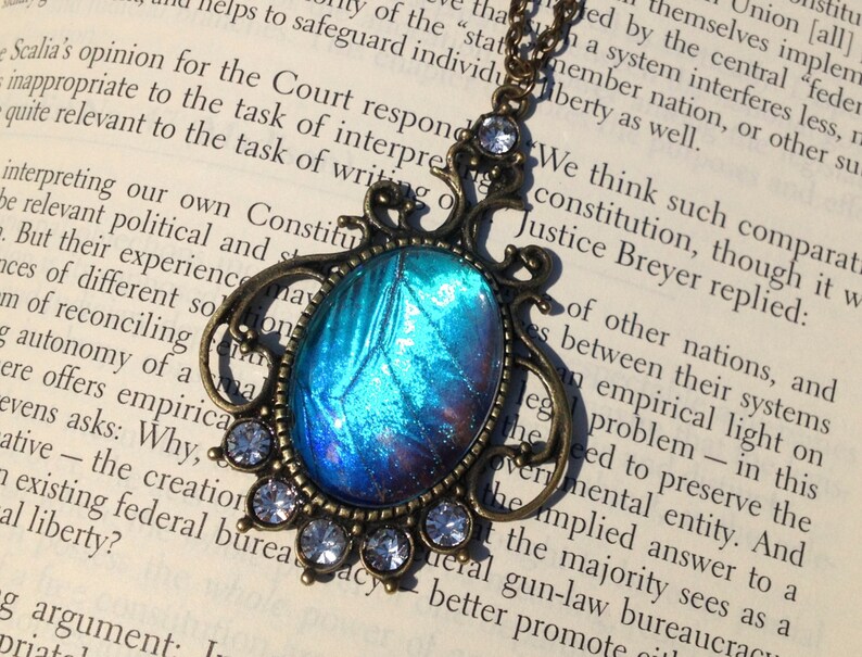 Puede incluir: Un collar de cadena de bronce con un colgante de dise&ntilde;o de ala de mariposa iridiscente azul y morado. El colgante est&aacute; rodeado de pedrer&iacute;a transparente.