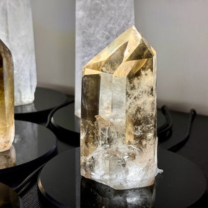 May include: A clear, faceted, golden citrine crystal point on a black display stand. The crystal is approximately 4 inches tall and has a natural, raw look.