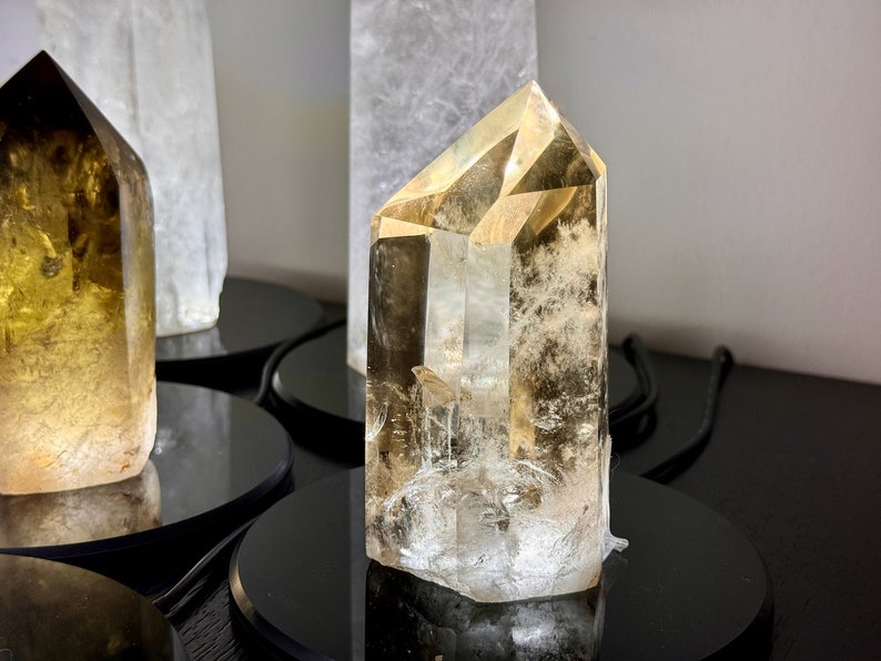 May include: A clear, faceted, golden citrine crystal point on a black display stand. The crystal is approximately 4 inches tall and has a natural, raw look.