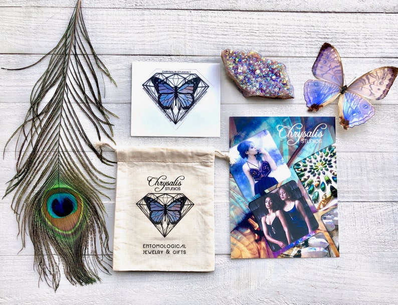 May include: A white canvas bag with the text "Chrysalis Studios" and "Entomological Jewelry & Gifts" printed on it. The bag features a blue and purple butterfly inside a diamond shape. The bag is sitting on a white wooden surface with a peacock feather, a crystal, and a butterfly sticker.