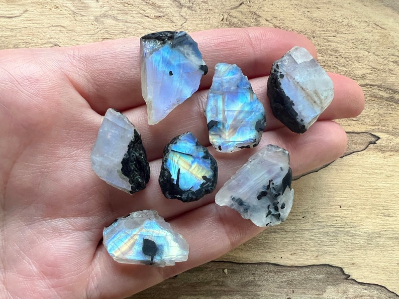 May include: A collection of seven iridescent rainbow moonstone rough crystals with black inclusions. The crystals are various shapes and sizes and have a white, translucent appearance.