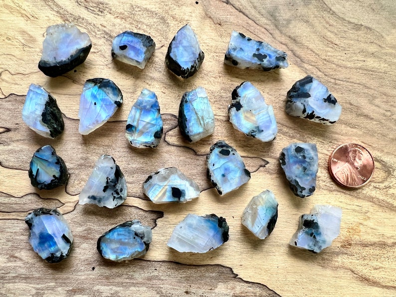 May include: A collection of 20 iridescent rainbow moonstone rough stones with black inclusions. The stones are various shapes and sizes and are arranged on a wooden surface. A penny is included for scale.