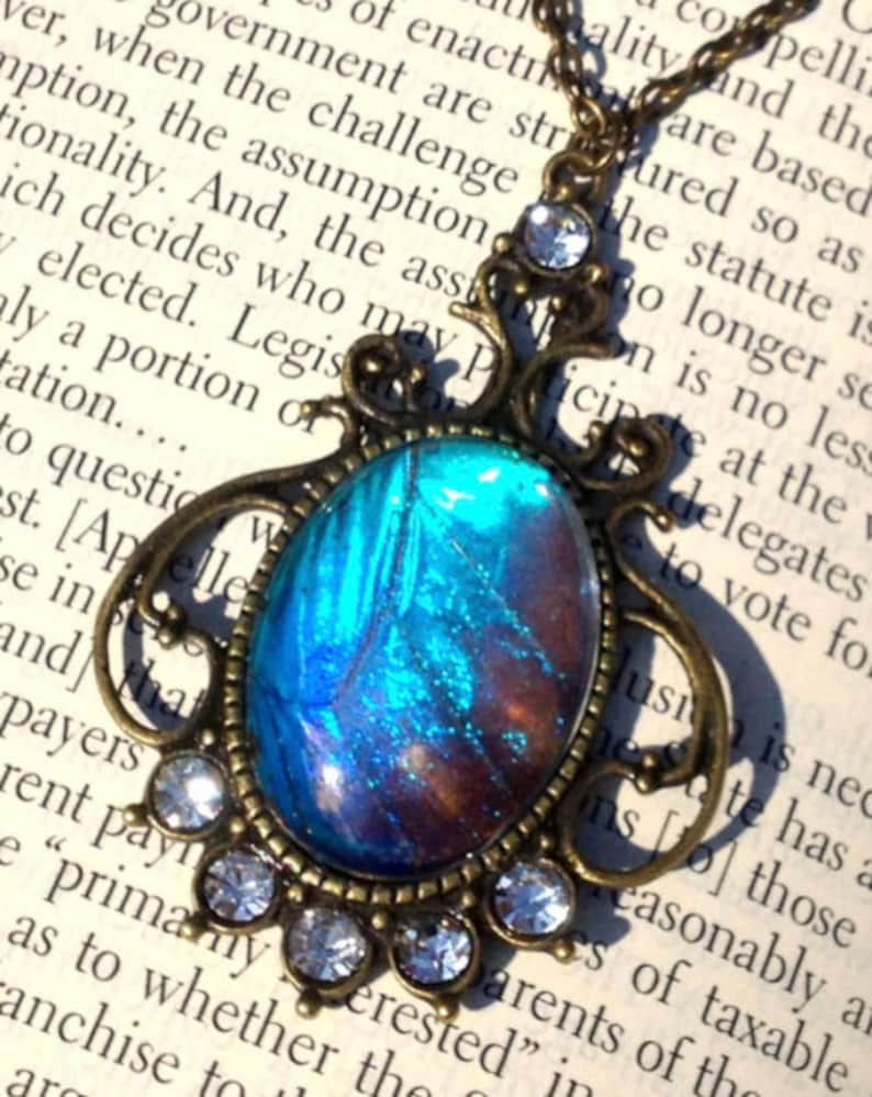Puede incluir: Un collar con un colgante de bronce con un ala de mariposa azul y marr&oacute;n encapsulada en resina. El colgante est&aacute; rodeado de diamantes de imitaci&oacute;n transparentes.