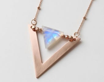 May include: A rose gold-toned necklace featuring a large, open triangular pendant with a brushed metal finish. Suspended within the centre of the triangle is a smaller, translucent, triangular moonstone with a blue iridescent sheen.