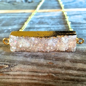 May include: A gold chain necklace with a white iridescent crystal bar pendant. The crystal bar is edged in gold.