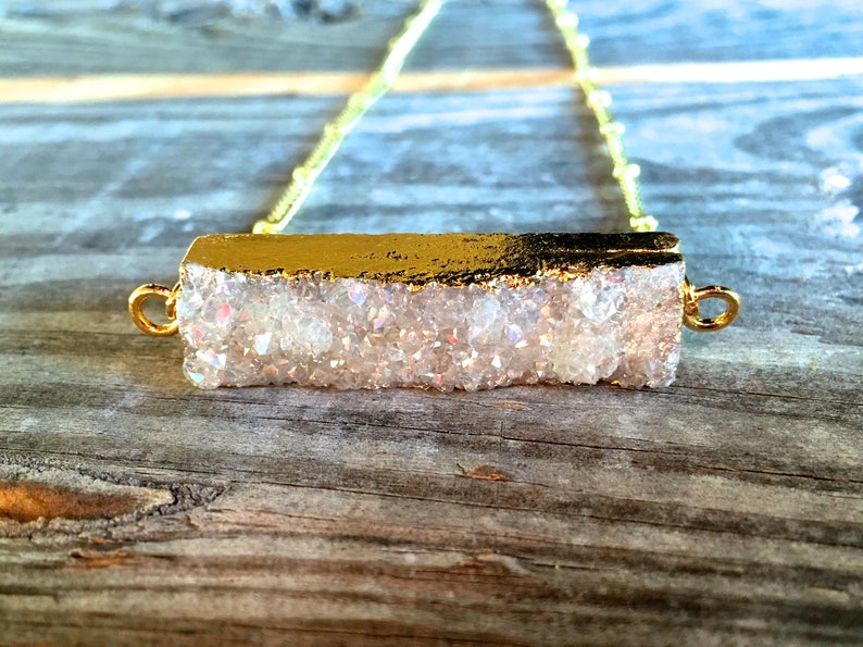 May include: A gold chain necklace with a white iridescent crystal bar pendant. The crystal bar is edged in gold.