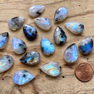 Puede incluir: Una colecci&oacute;n de 15 cabujones de piedra lunar arco&iacute;ris en forma de l&aacute;grima con un brillo nacarado. Las piedras son una mezcla de blanco, azul y gris con inclusiones negras. Las piedras est&aacute;n dispuestas sobre una superficie de madera.