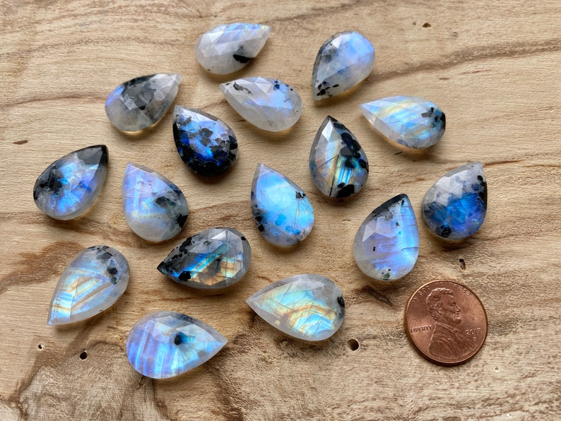 Puede incluir: Una colecci&oacute;n de 15 cabujones de piedra lunar arco&iacute;ris en forma de l&aacute;grima con un brillo nacarado. Las piedras son una mezcla de blanco, azul y gris con inclusiones negras. Las piedras est&aacute;n dispuestas sobre una superficie de madera.