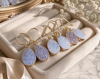 Puede incluir: Tres pares de pendientes en forma de lágrima con ganchos dorados. Cada pendiente presenta una superficie de piedra druzy brillante e iridiscente, engastada en un marco de metal dorado, dispuestos ordenadamente en un joyero de terciopelo beige.