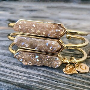 May include: Three gold bangles with a light brown crystal-like material on the front. The bangles are stacked on top of each other. A gold heart charm and a gold charm with the letter "C" are attached to the bottom bangle.