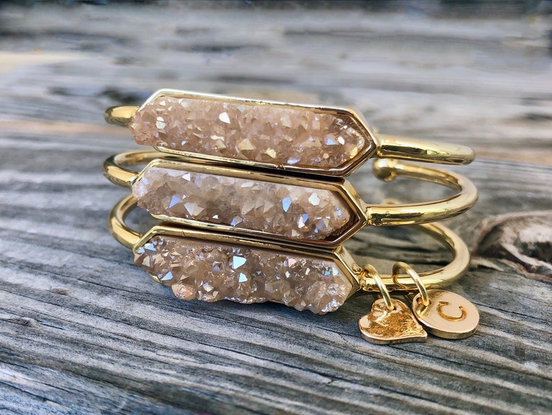 May include: Three gold bangles with a light brown crystal-like material on the front. The bangles are stacked on top of each other. A gold heart charm and a gold charm with the letter "C" are attached to the bottom bangle.