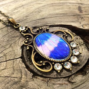Op de afbeelding: Een vintage-stijl hangerketting met een grote, ovaalvormige blauw-paarse iriserende steen. De steen is gezet in een sierlijk, antiek goudkleurig metalen frame, geaccentueerd met heldere kristallen. De ketting hangt aan een delicate ketting.
