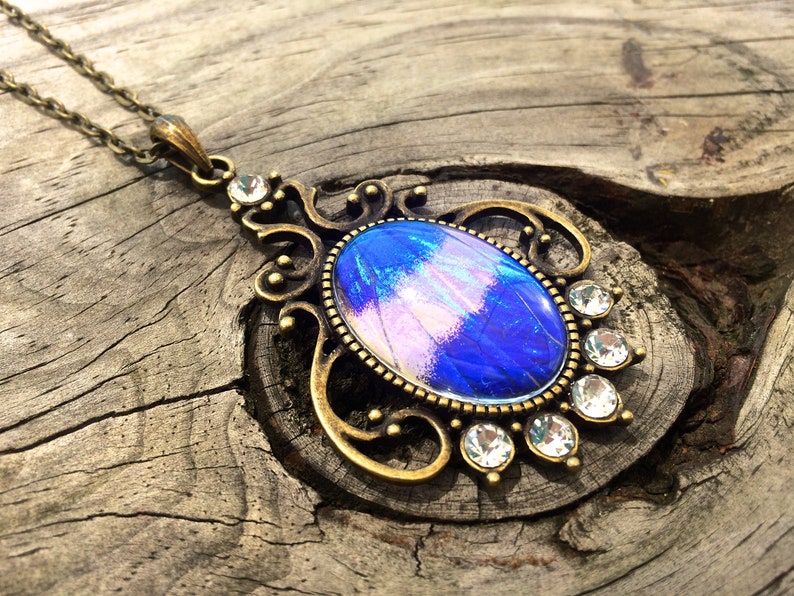 Op de afbeelding: Een vintage-stijl hangerketting met een grote, ovaalvormige blauw-paarse iriserende steen. De steen is gezet in een sierlijk, antiek goudkleurig metalen frame, geaccentueerd met heldere kristallen. De ketting hangt aan een delicate ketting.