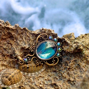 May include: A bronze pendant necklace featuring a blue and green iridescent glass cabochon. The pendant is embellished with small, clear rhinestones. The necklace is pictured on a rocky surface.