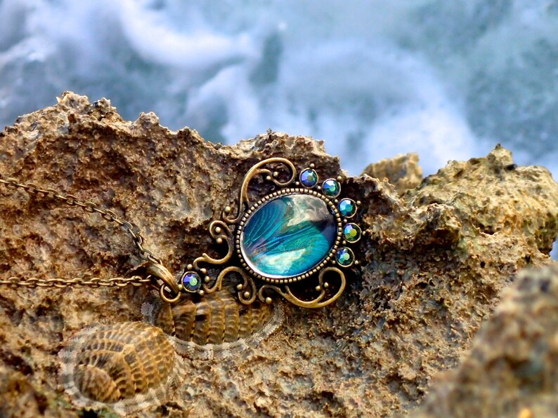 May include: A bronze pendant necklace featuring a blue and green iridescent glass cabochon. The pendant is embellished with small, clear rhinestones. The necklace is pictured on a rocky surface.