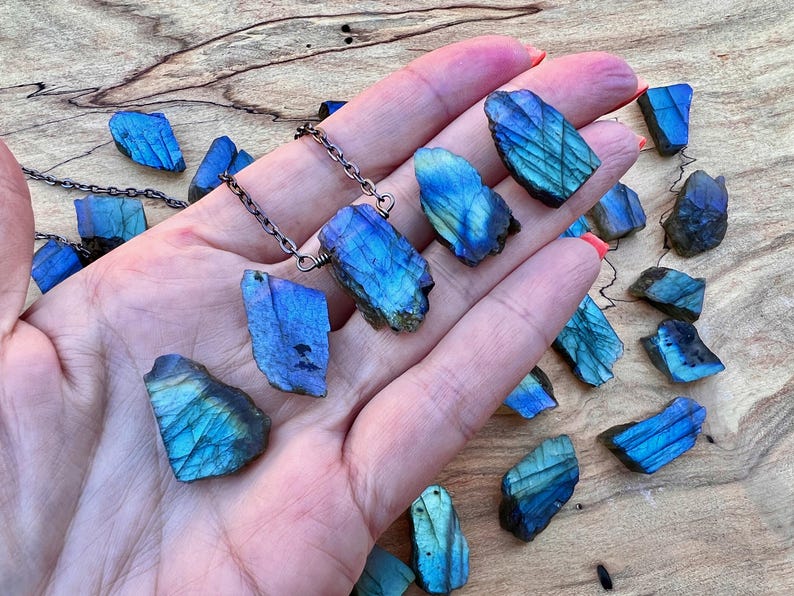 A hand holding a necklace with blue and green stones, as well as some additional blue and green stones scattered on a wooden surface.