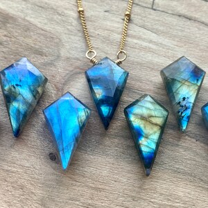 May include: Seven iridescent blue labradorite gemstone pendants in a kite shape, with a gold chain necklace in the background.