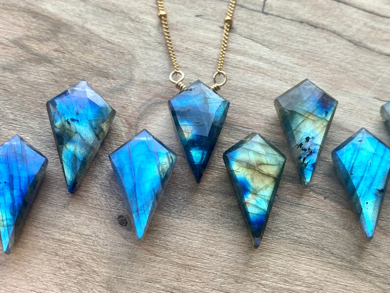 May include: Seven iridescent blue labradorite gemstone pendants in a kite shape, with a gold chain necklace in the background.