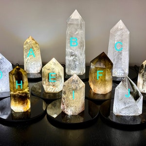 May include: Ten clear quartz crystal points with various shades of yellow and green, displayed on black circular bases. The crystals are labeled A through J.
