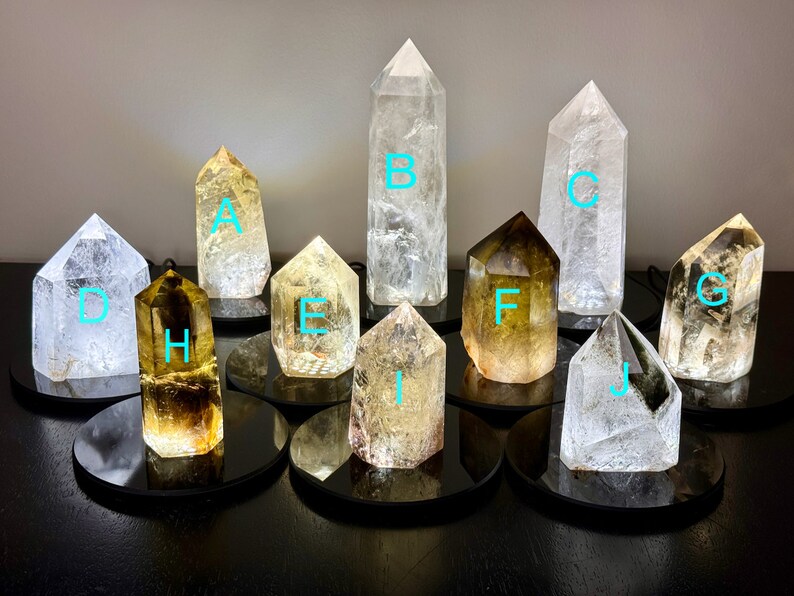 May include: Ten clear quartz crystal points with various shades of yellow and green, displayed on black circular bases. The crystals are labeled A through J.