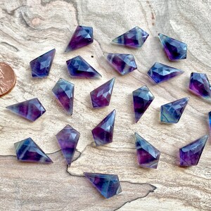 May include: A collection of 20 purple and blue fluorite points, each with a faceted, diamond-shaped design. The points are arranged in a random pattern on a wooden surface.