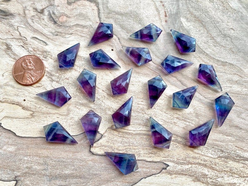 May include: A collection of 20 purple and blue fluorite points, each with a faceted, diamond-shaped design. The points are arranged in a random pattern on a wooden surface.