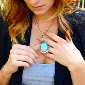 A woman wearing a necklace with a pendant is looking down at her necklace, which has a blue and green stone.