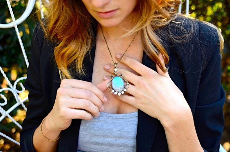 A woman wearing a necklace with a pendant is looking down at her necklace, which has a blue and green stone.