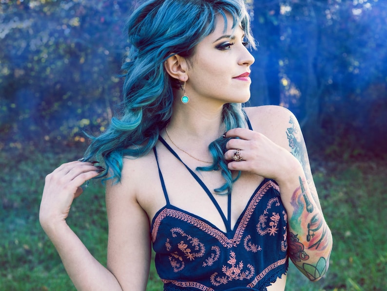 May include: A woman with blue hair wearing a blue and pink floral crop top with a black strap. She has a tattoo on her arm and is wearing a ring on her finger.