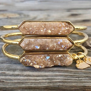 May include: Three gold bangles with a light brown crystal charm. The charms are in the shape of a rectangle with rounded edges. The bangles are stacked on top of each other. The charm on the bottom bangle has a small gold heart charm attached to it. The charm on the top bangle has a small gold charm with the letter "G" on it.
