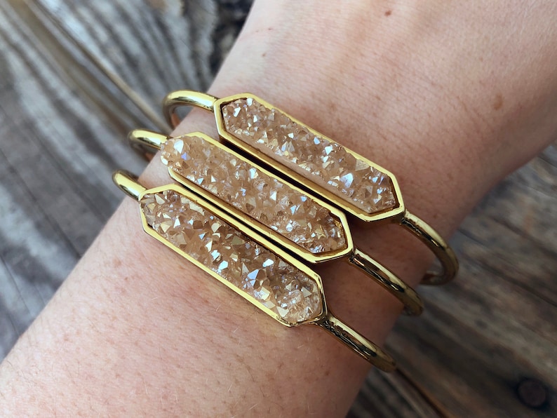 May include: Three gold bangles with a faceted, light brown crystal bar on each. The bangles are stacked on a person's wrist.