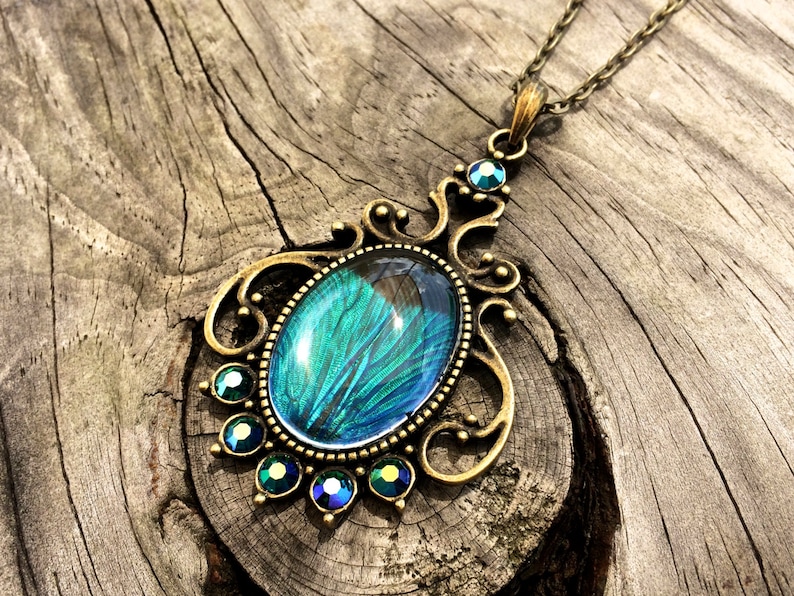 May include: A bronze pendant necklace featuring a large oval-shaped blue and green gemstone set in a filigree frame. The gemstone has a peacock feather design.