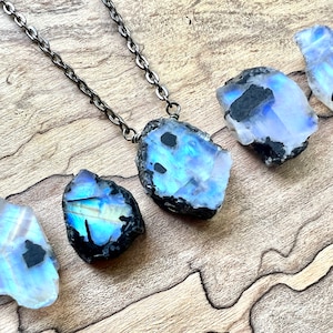 May include: A silver chain necklace with a large, iridescent moonstone pendant. The moonstone is raw and has a black, rough exterior. The necklace is displayed on a wooden surface.