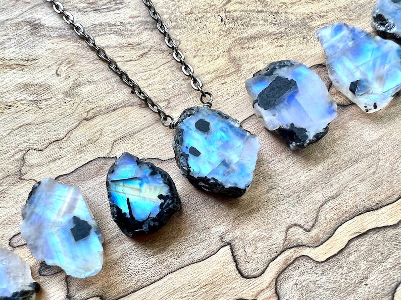 May include: A silver chain necklace with a large, iridescent moonstone pendant. The moonstone is raw and has a black, rough exterior. The necklace is displayed on a wooden surface.