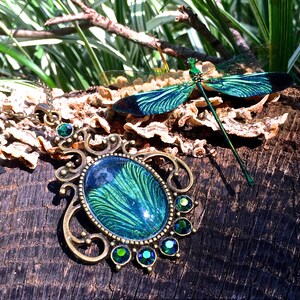 May include: A green and blue dragonfly pendant necklace with a detailed ornate design and green crystal accents. The necklace is on a wooden surface with a dragonfly brooch nearby.