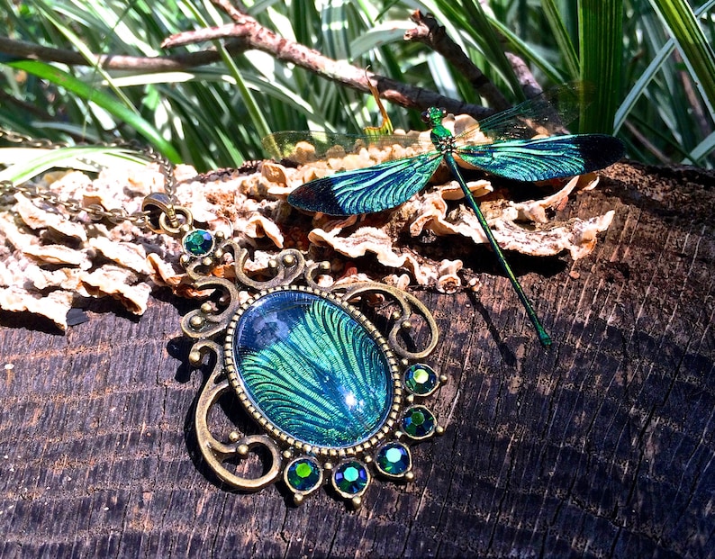 May include: A green and blue dragonfly pendant necklace with a detailed ornate design and green crystal accents. The necklace is on a wooden surface with a dragonfly brooch nearby.