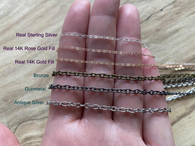 A hand holding various types of chain necklaces, including Sterling Silver, 14K Rose Gold Fill, Bronze, Gunmetal, and Antique Silver.
